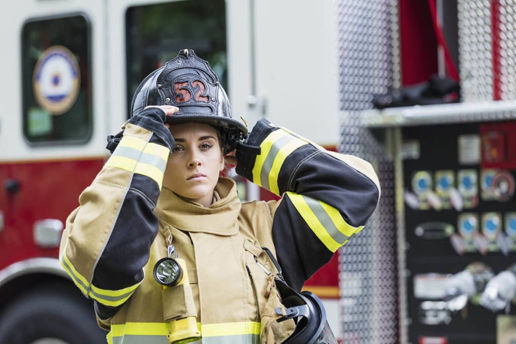 A female firefighter wearing a fire protection suit, standing next to a fire engine. She is a young woman in her 20s with a serious expression.