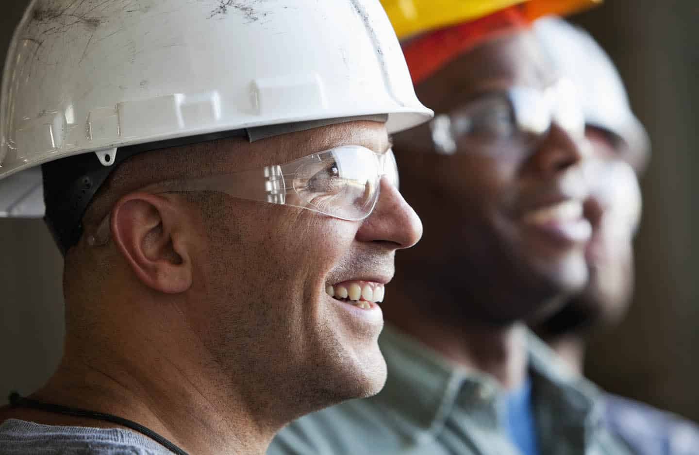 Close up of group of multi-ethnic construction workers wearing hard hats and safety glasses.  Focus on man in foreground (30s).