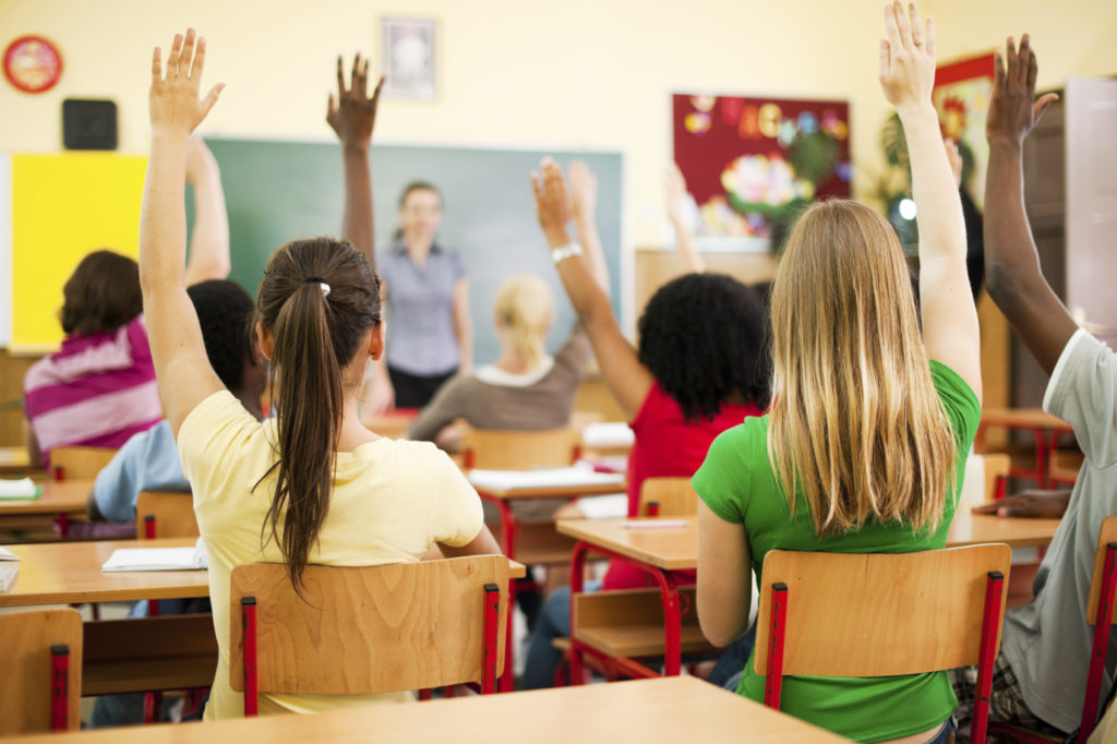 Group of teenagers sitting in classroom with raised hands