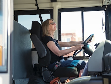 Portrait of mature woman driving a school bus. She ha her security belt on and two hands on the wheel. Horizontal waist up outdoors shot on a bright sunny day. Copy space.