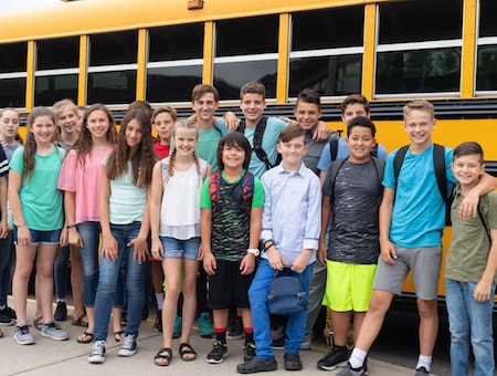 A multiethnic group of elementary students pose and goof around for a picture outside the school bus. Some are looking at the camera.