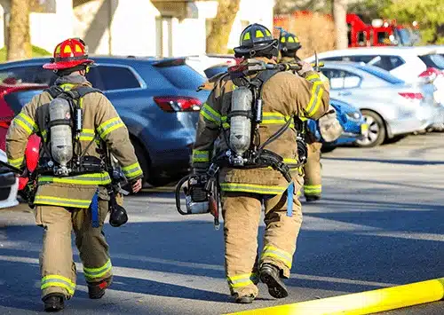 A group of firefighters in full turnout gear and SCBA walk down a street lined with cars holding equipment in the daytime.
