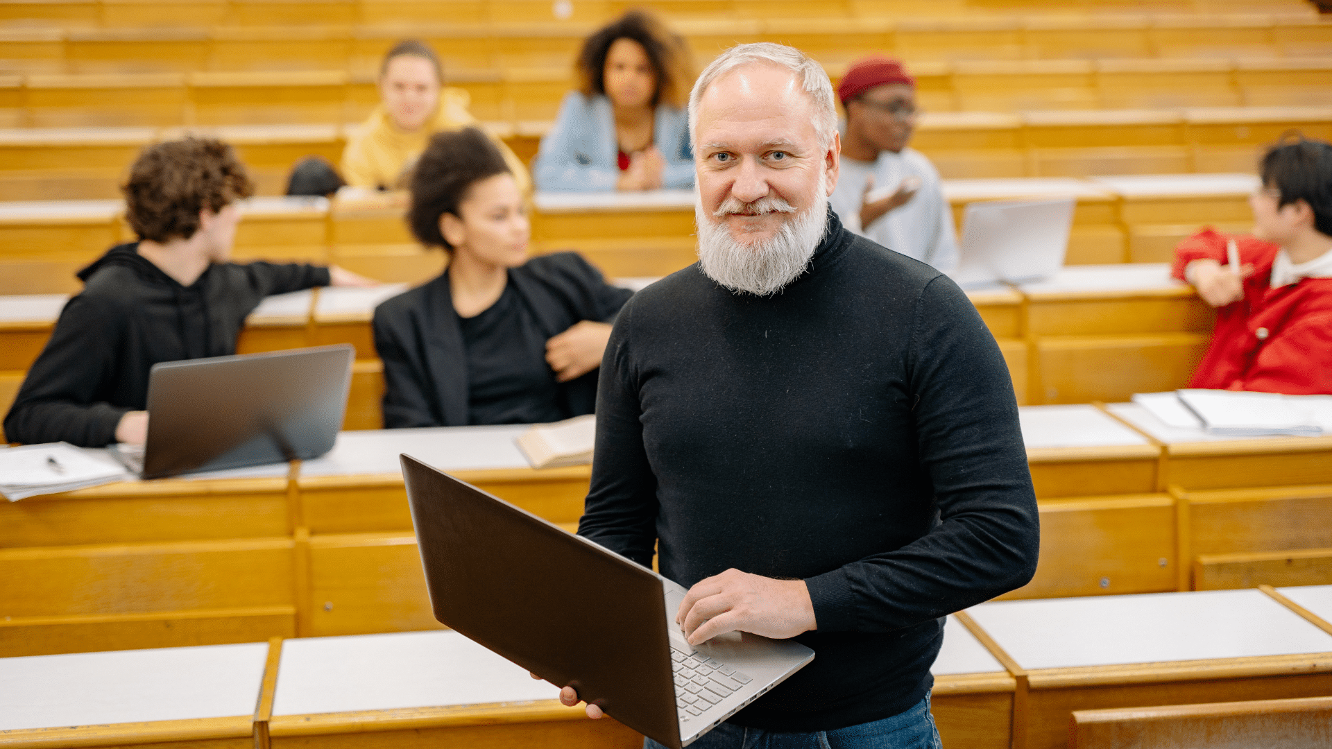 A professor standing in front of a lecture hall holding a computer
