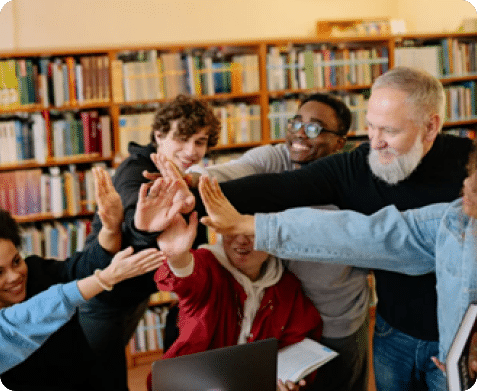 A group of students and a teacher in a library reaching in for a team high-five around a laptop.