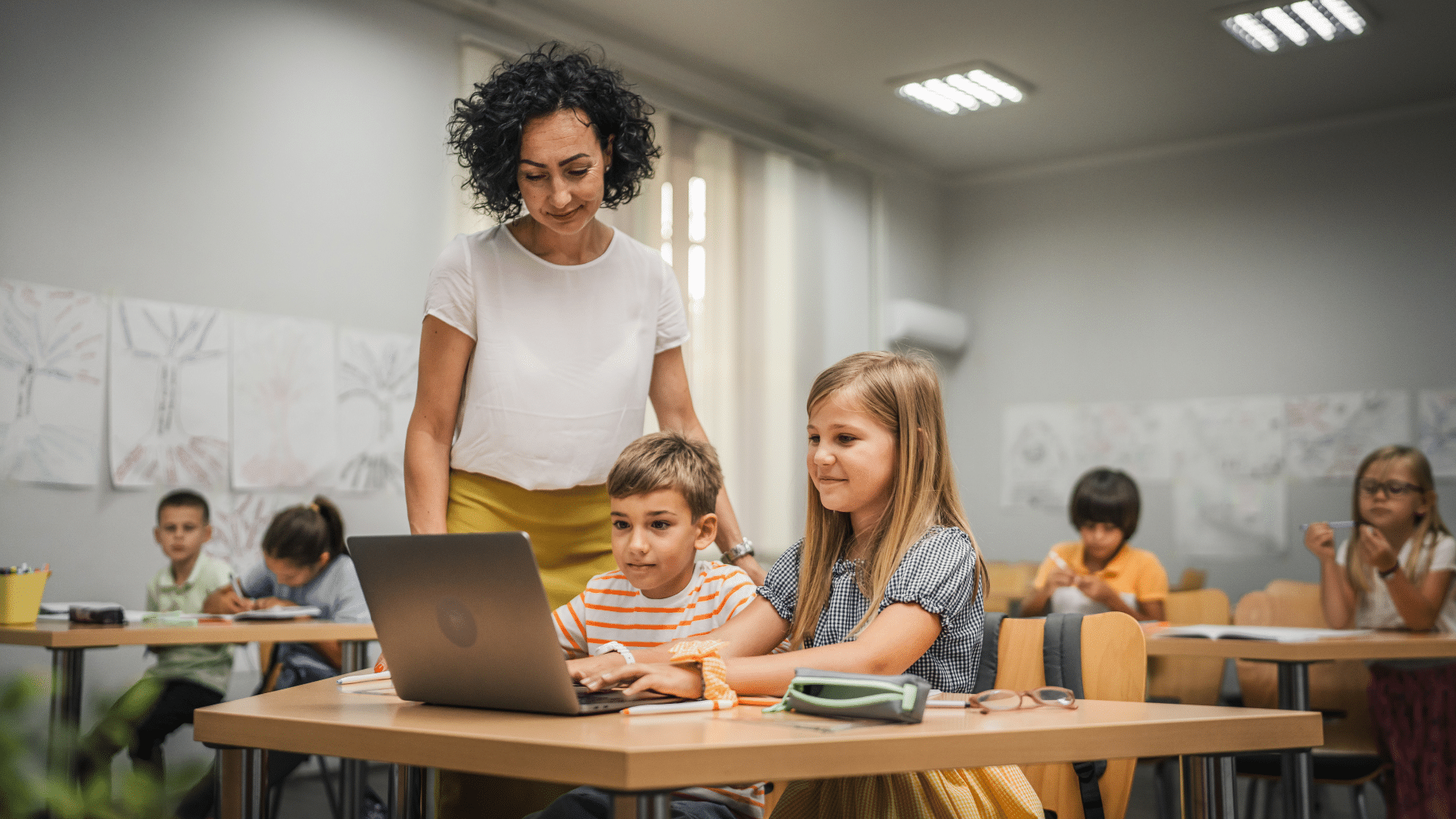 Two young students using a computer with a teacher overlooking them.