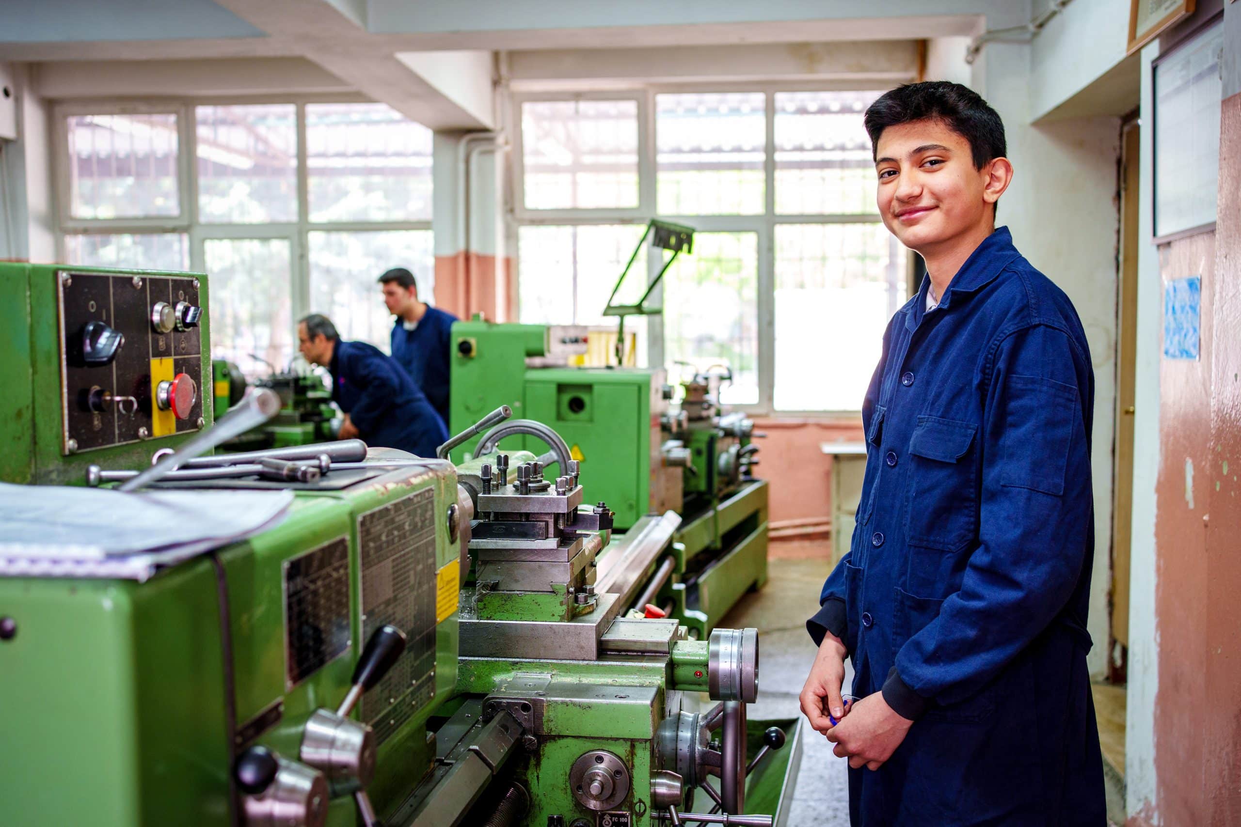 A young male trainee in safety glasses operates a Lathe machine in a technical training workshop.