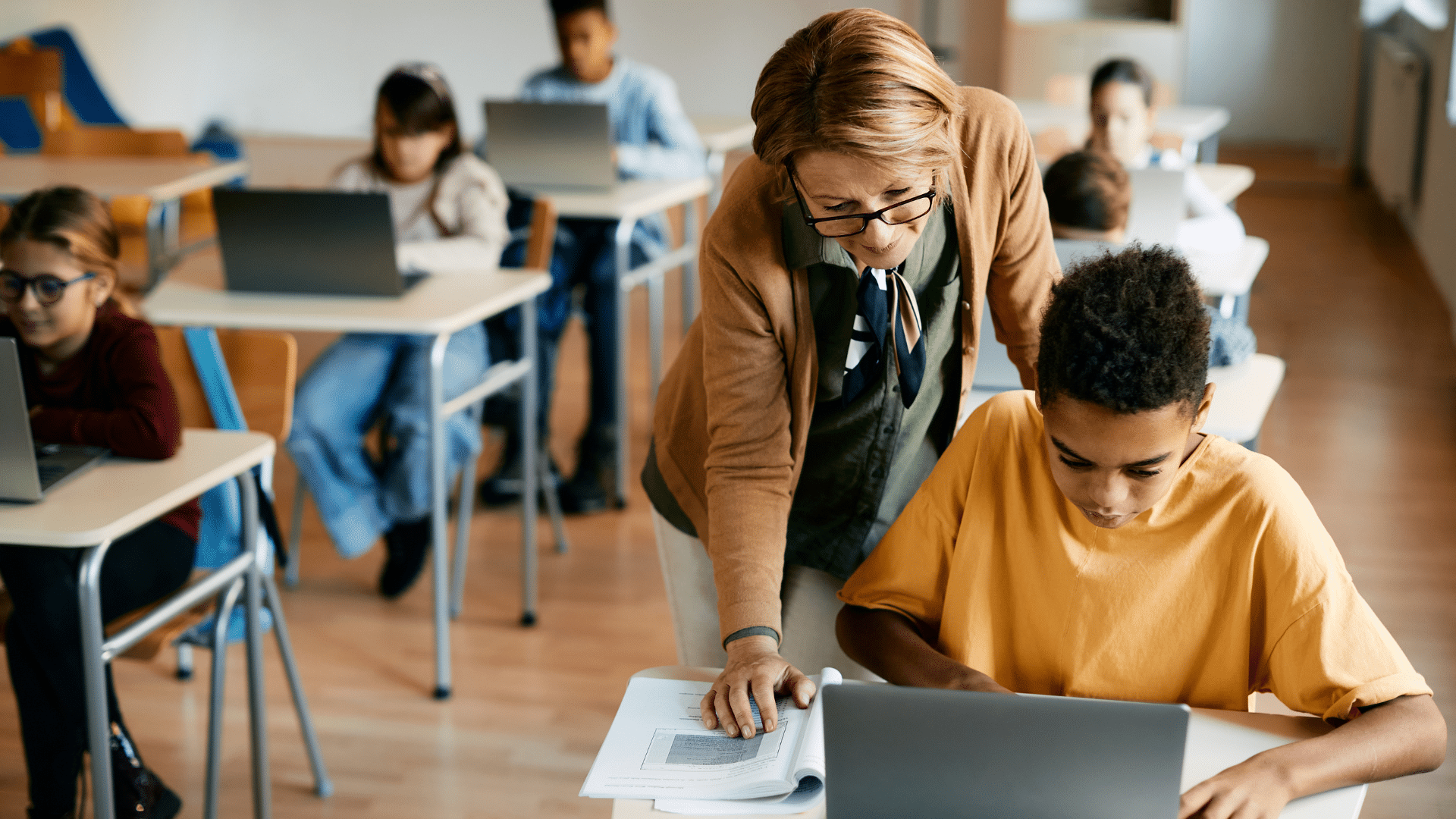A K-12 teacher assisting a student working on a laptop.