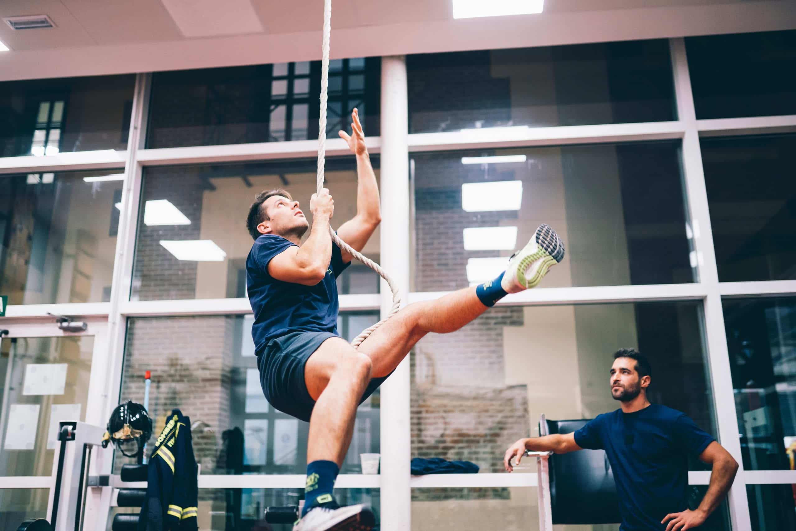 Volunteer firefighter fitting in training at the station between calls, climbing a rope as a crewmate watches.